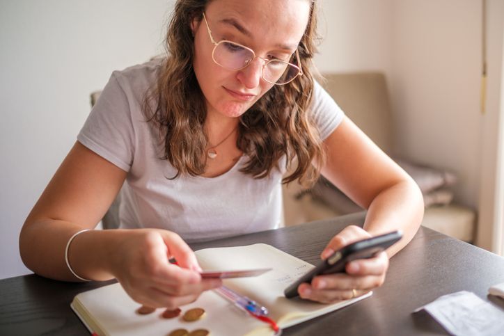 Person sitting at a table, checking a smartphone while writing notes in a notebook.