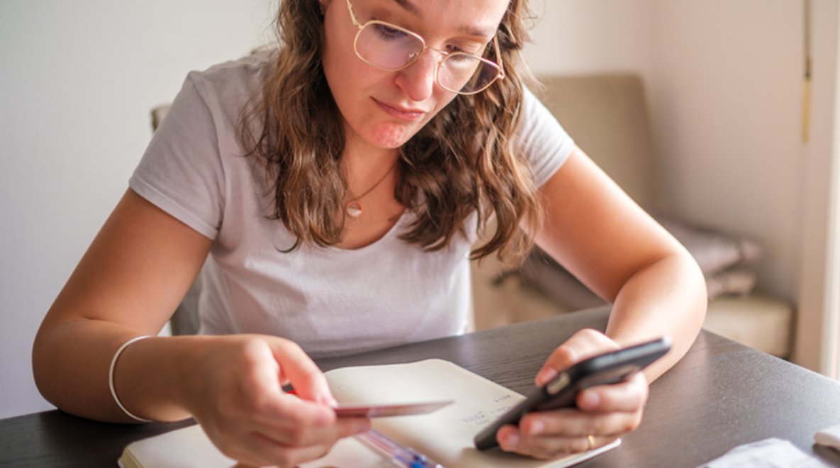Person sitting at a table, checking a smartphone while writing notes in a notebook.